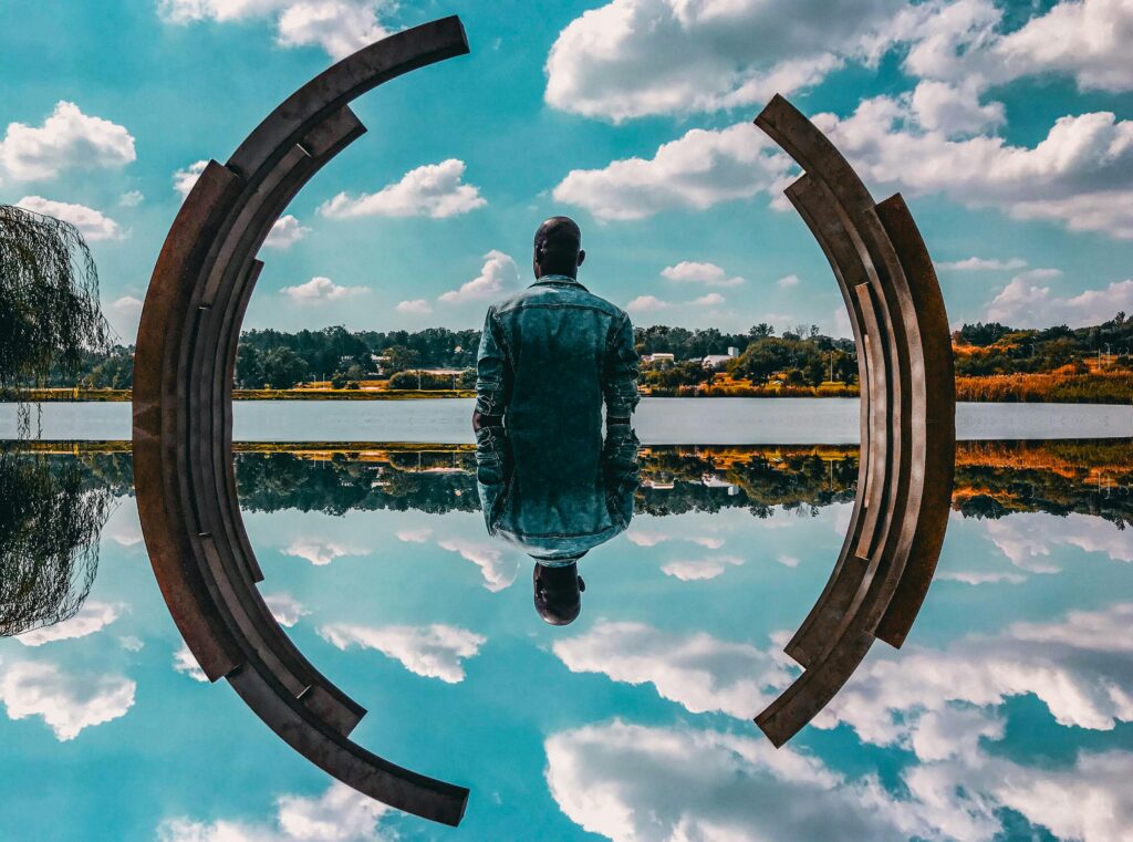 A man in a denim jacket sits by abstract sculpture reflecting on the water under a blue sky.